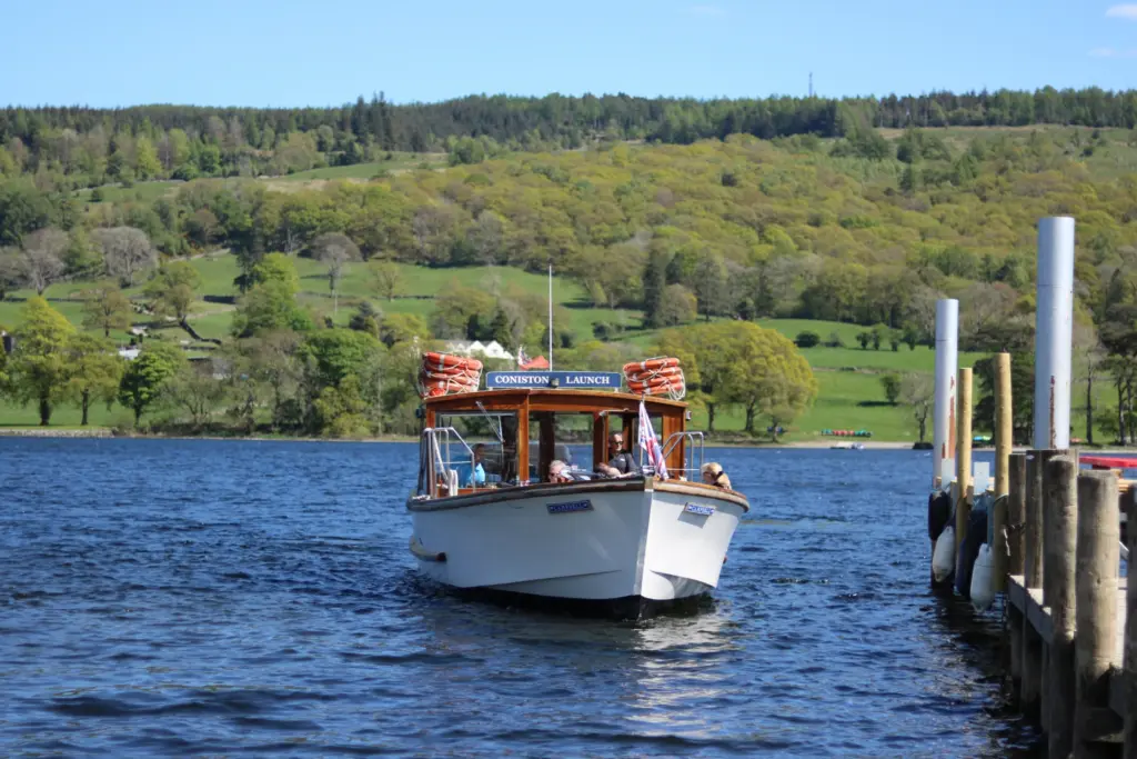 Coniston Launch boat carrying passengers across Coniston Water on a sunny day, with woodland and fields in the back drop