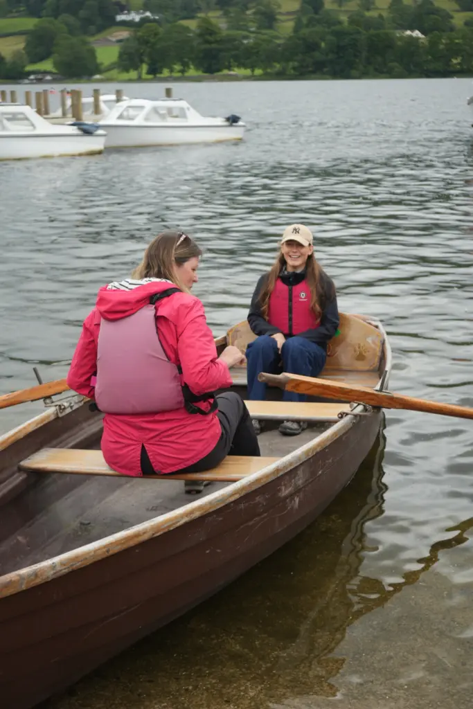 Two adult females on a wooden rowing boat with a wooden jetty and motor boats behind