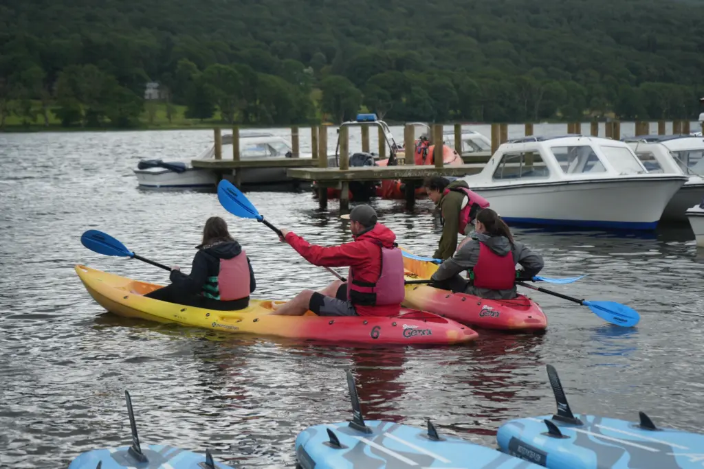 Four people on two double kayaks on the water in front of a jetty and boat