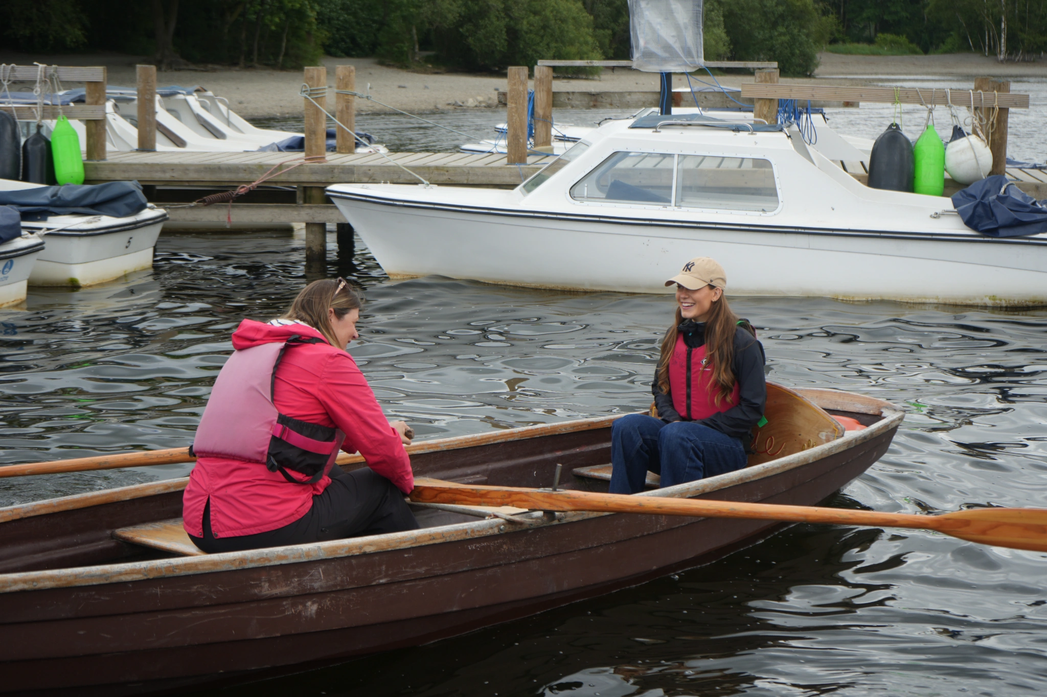 Two adult females on a wooden rowing boat with a wooden jetty and motor boats behind